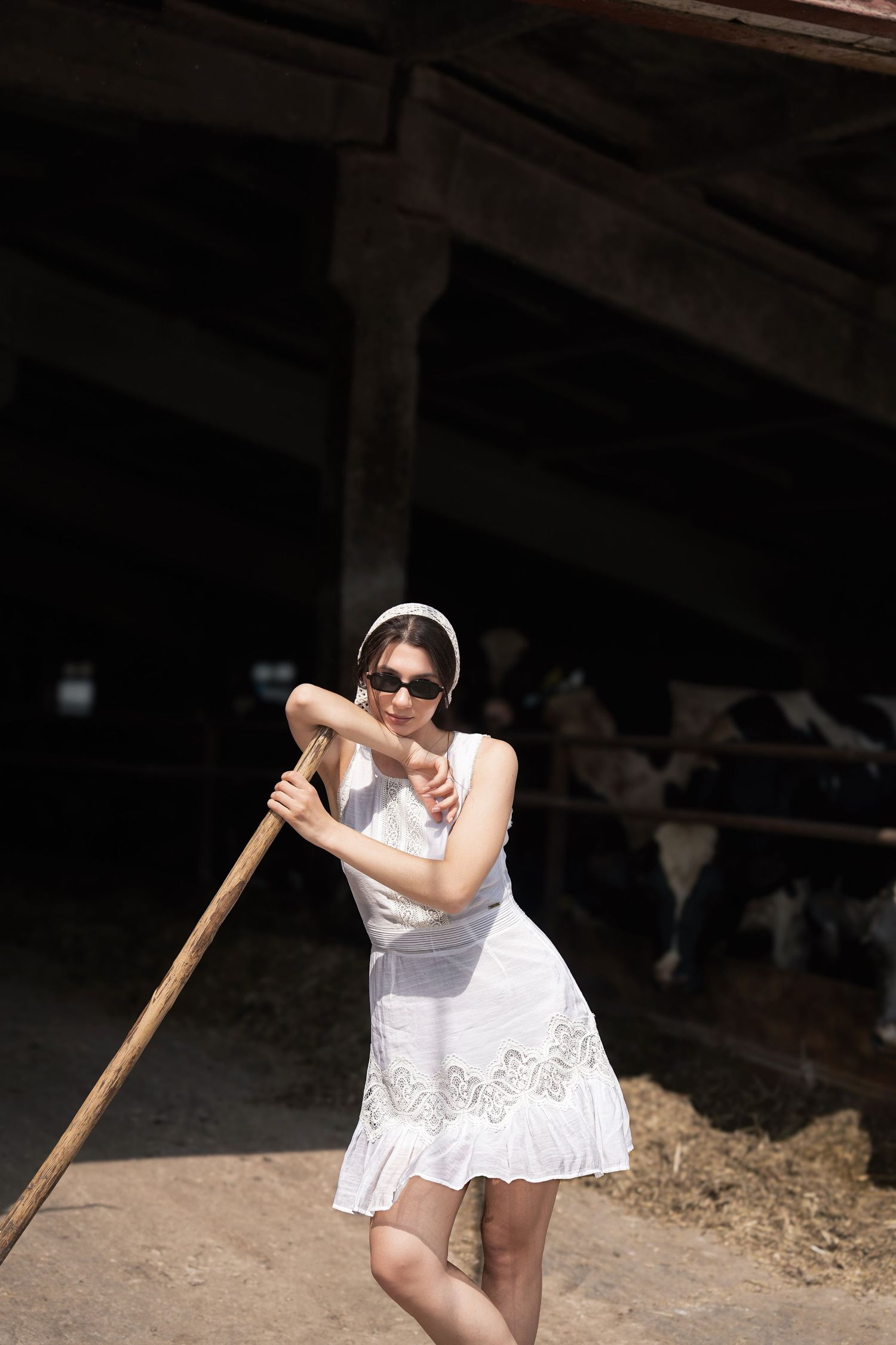 Young woman in white dress leaning on wooden staff in rustic barn with cows.