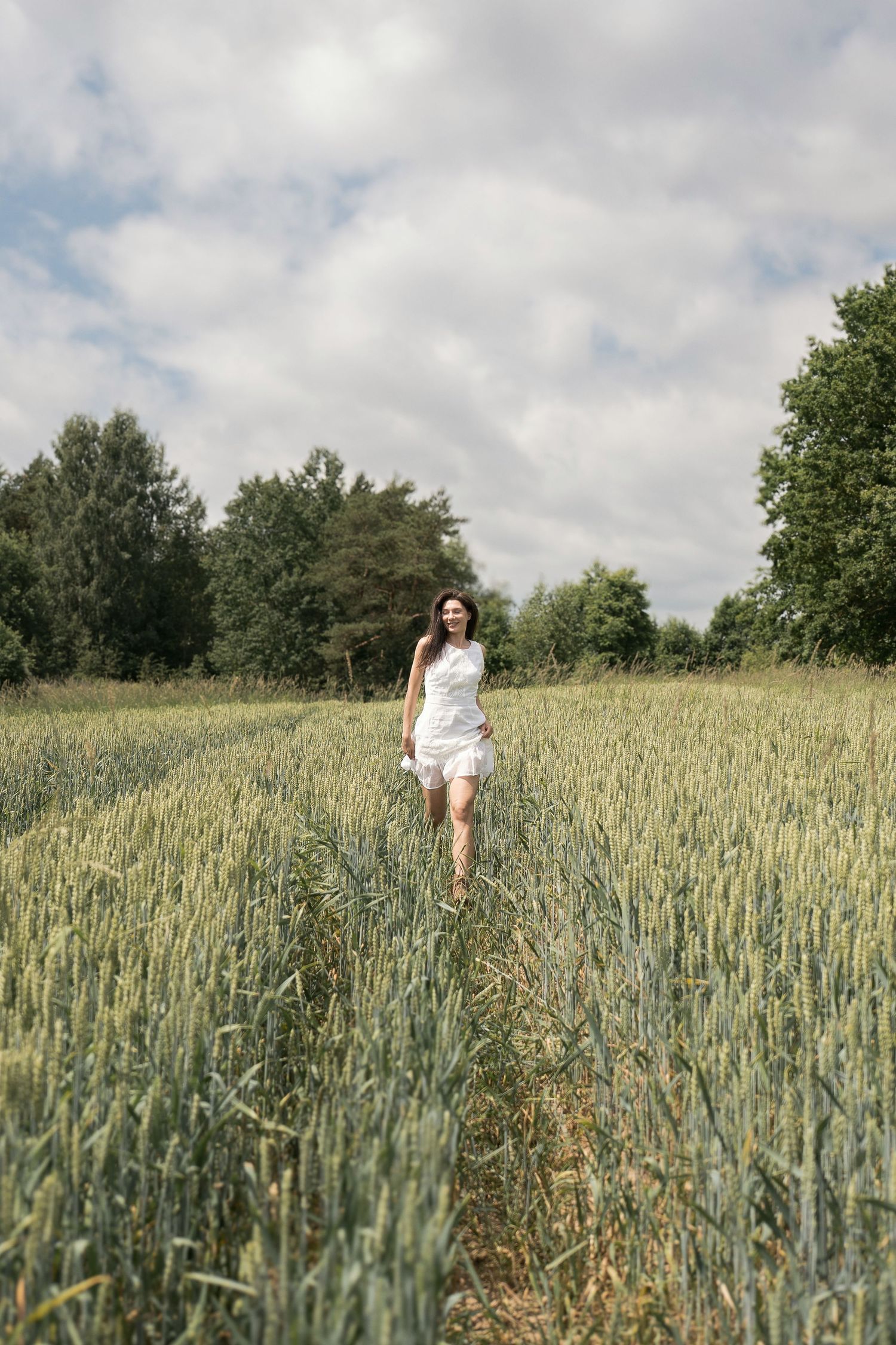 Young woman in white mini dress walking through a green wheat field under cloudy sky.