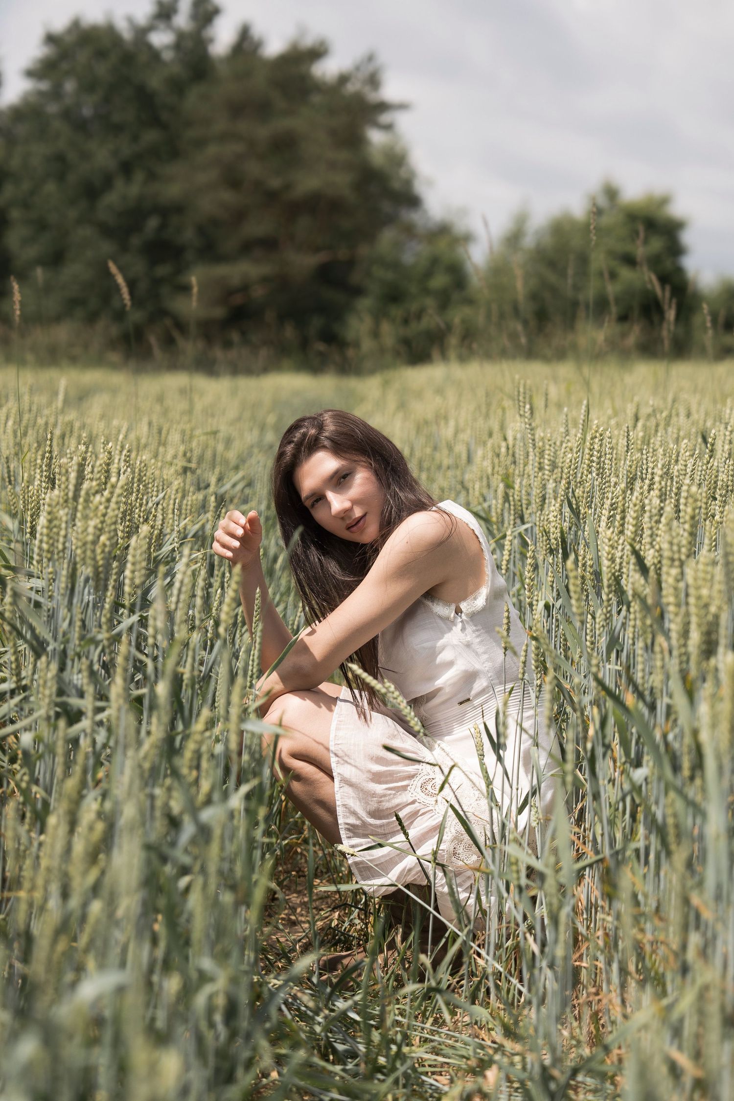 Young woman in white dress crouching in a green wheat field, holding grain stalks, with trees in the background.