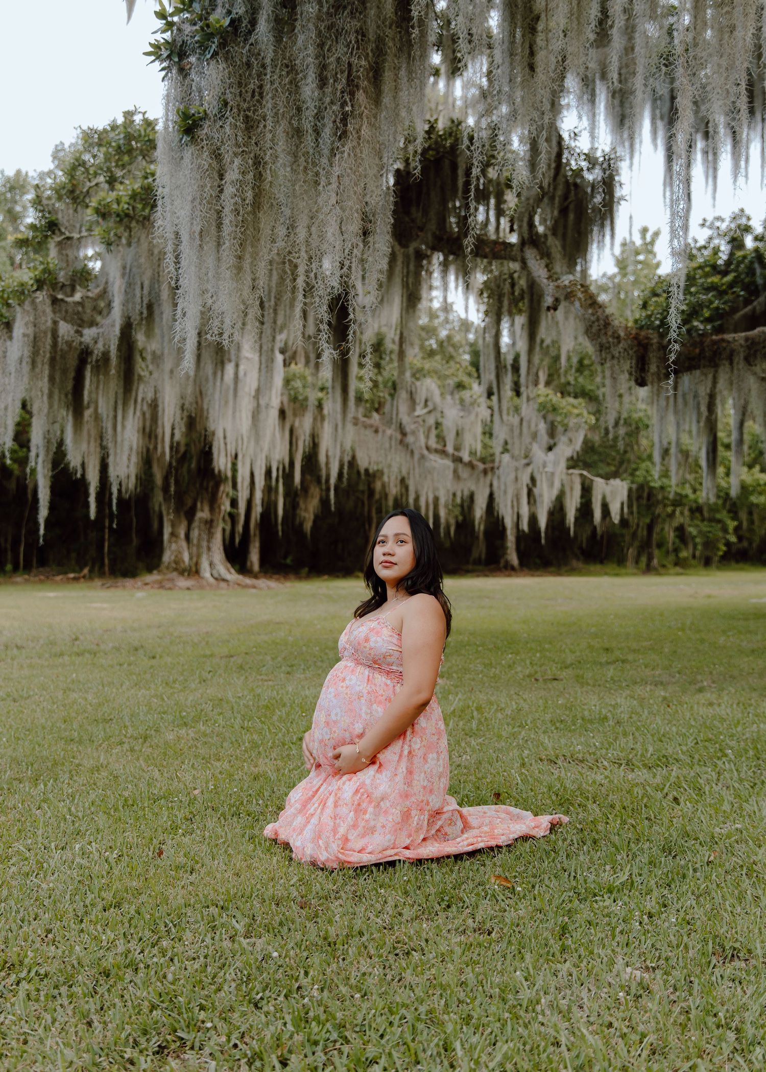 Pregnant woman in pink floral dress kneeling on grass under Spanish moss-draped oak trees during maternity photo session.