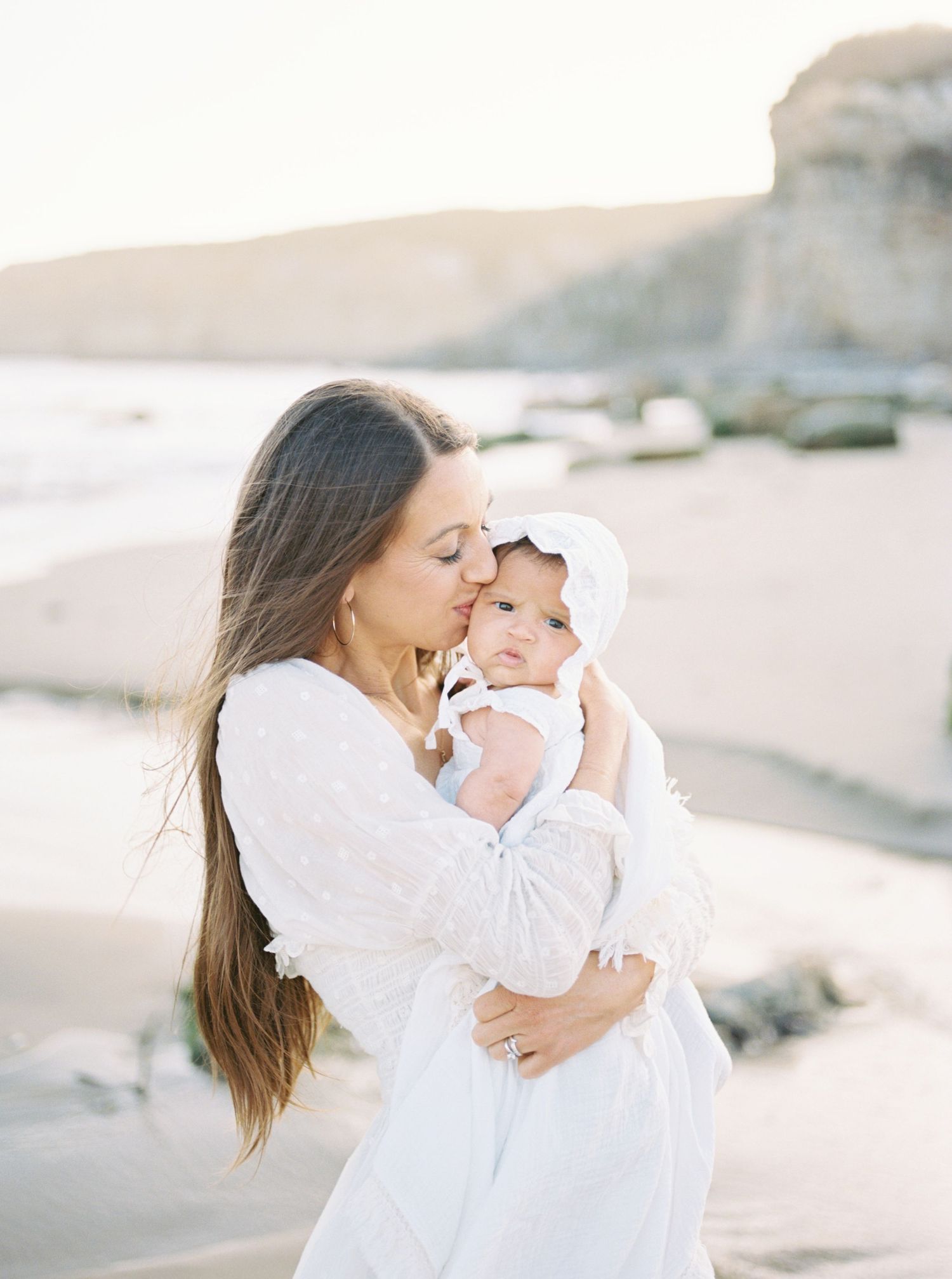 Mother in white dress kissing her baby on the beach at golden hour, both dressed in white.