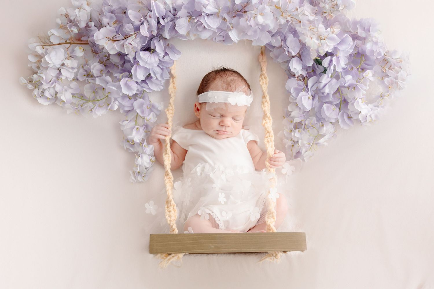 Newborn baby sleeping peacefully on a wooden swing adorned with purple wisteria flowers in a white romper and headband.