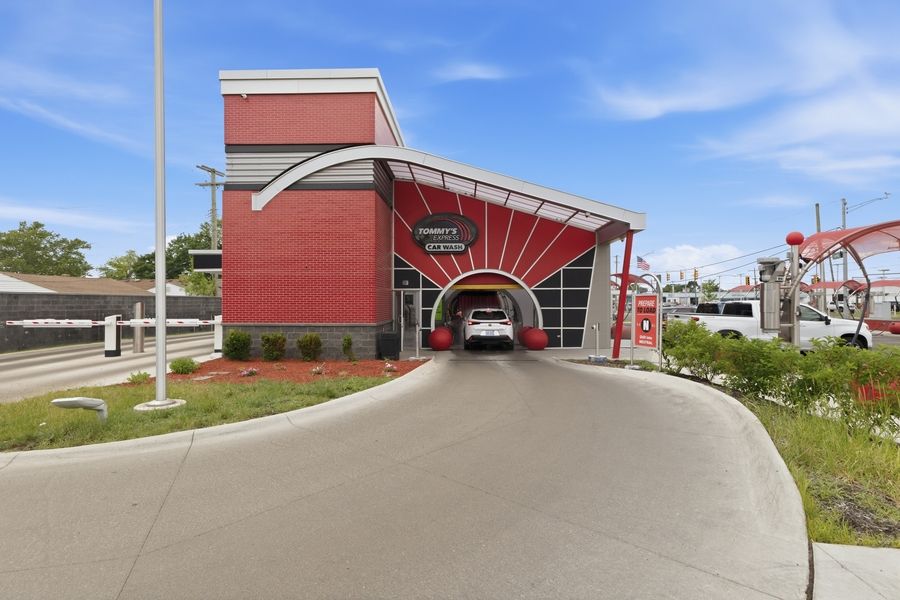 Red modern car wash building with drive-through tunnel entrance, curved canopy, and landscaped driveway under blue sky.