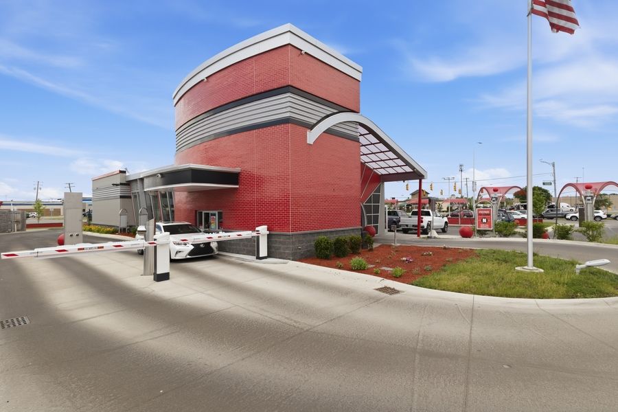 Modern red brick self-storage facility with security gate, canopy entrance, and American flag under clear blue sky.
