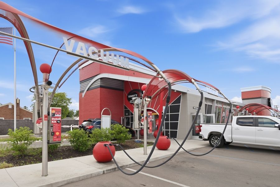 Exterior of a Macs convenience store with red decorative metal arches and globe sculptures in a parking lot under blue sky.