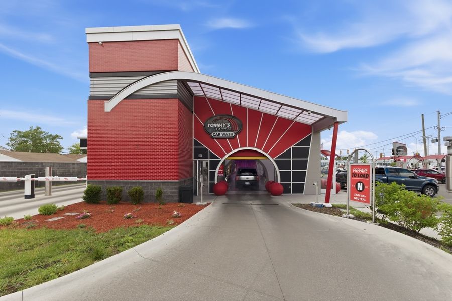 Red car wash building with tunnel entrance, canopy, and landscaping under blue sky.