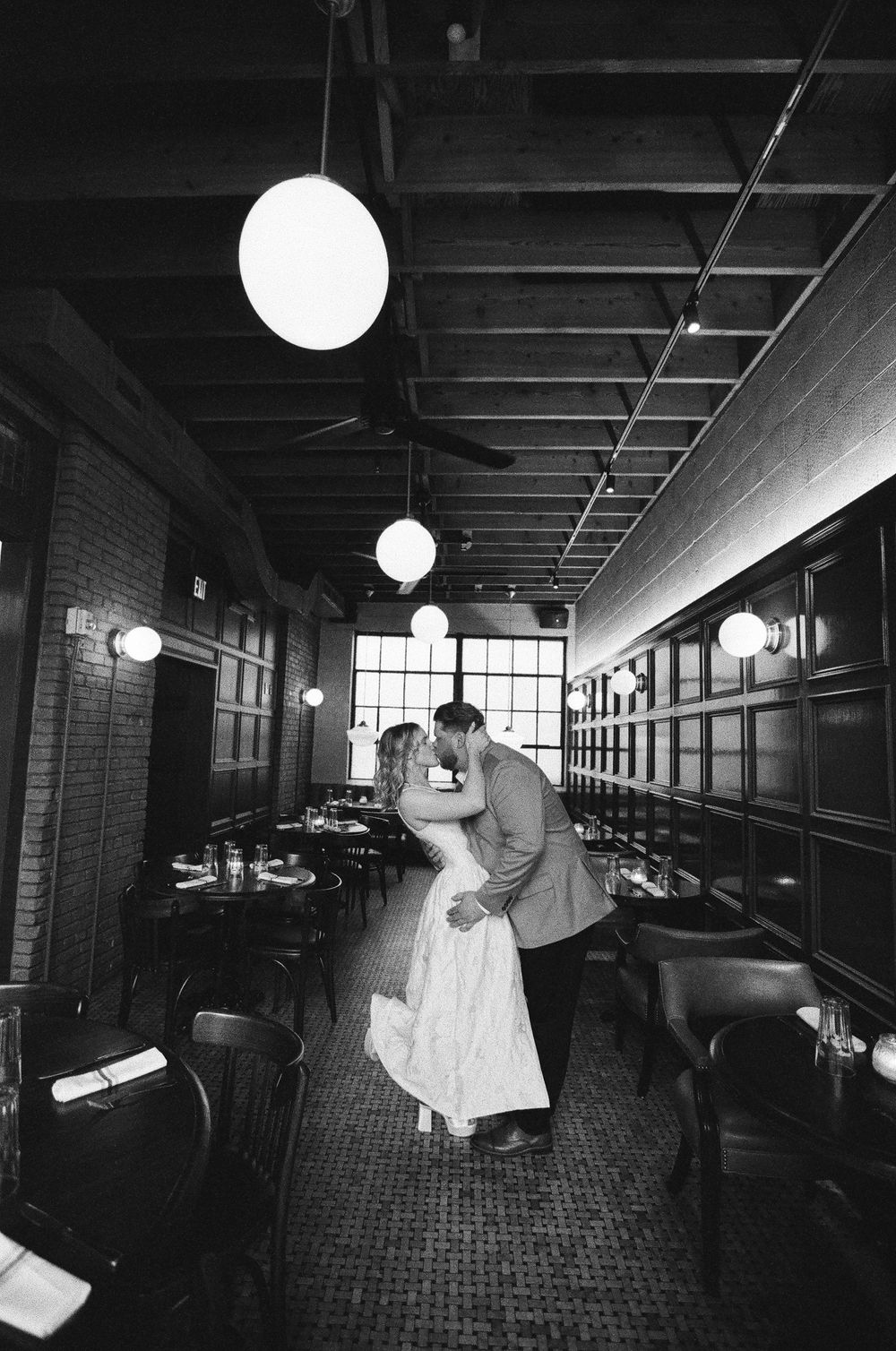 Black and white photo of a couple sharing a romantic kiss in an elegant restaurant with pendant lights and brick walls.