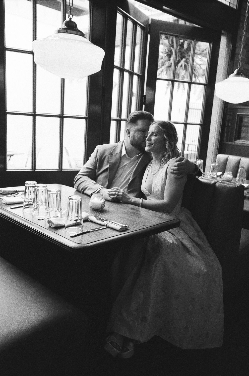 Black and white photo of a couple sitting together at a restaurant booth, smiling and embracing near large windows.
