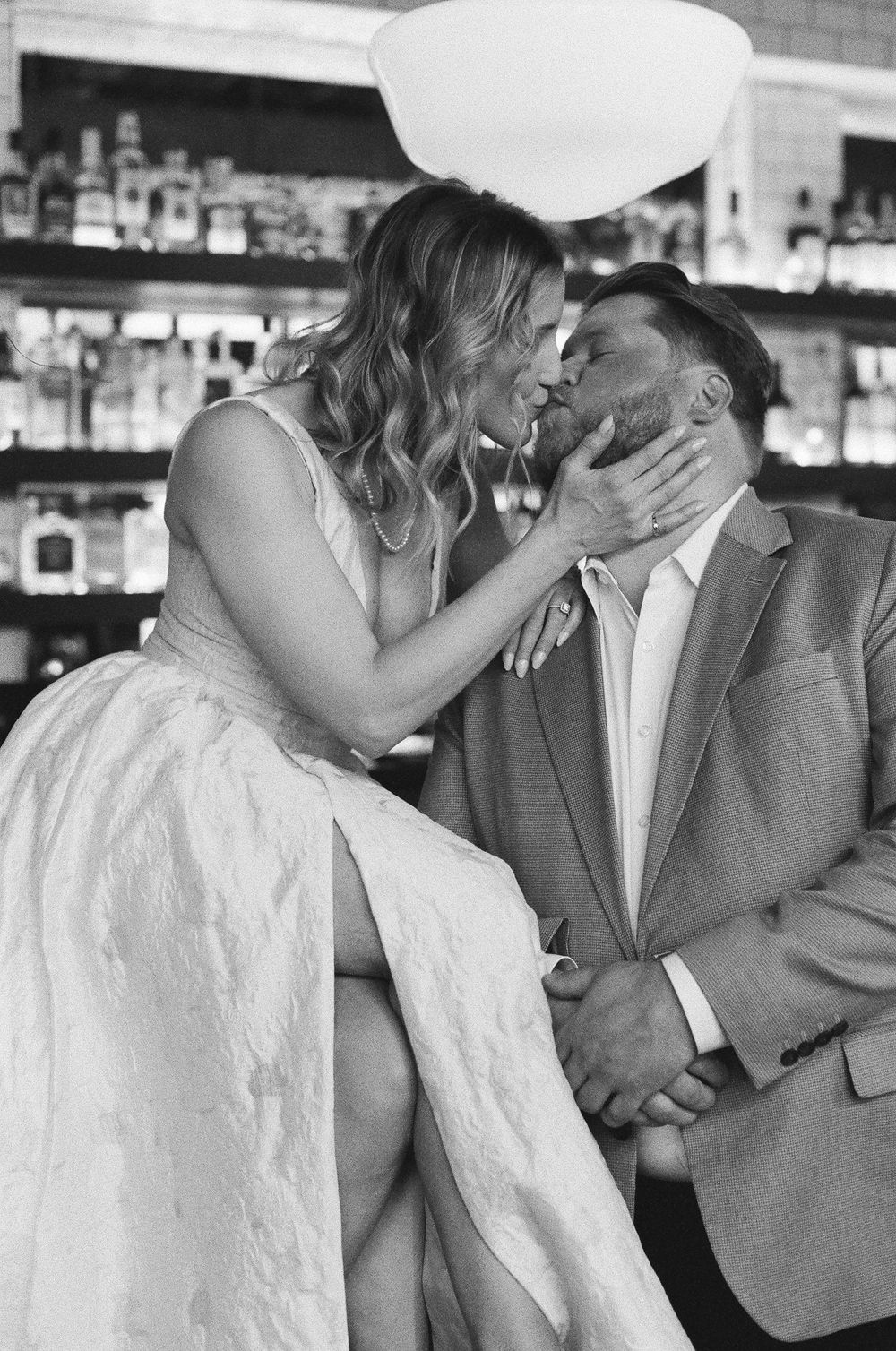 Black and white photo of a couple sharing a romantic kiss at a bar, bride in white dress holding groom's face tenderly.