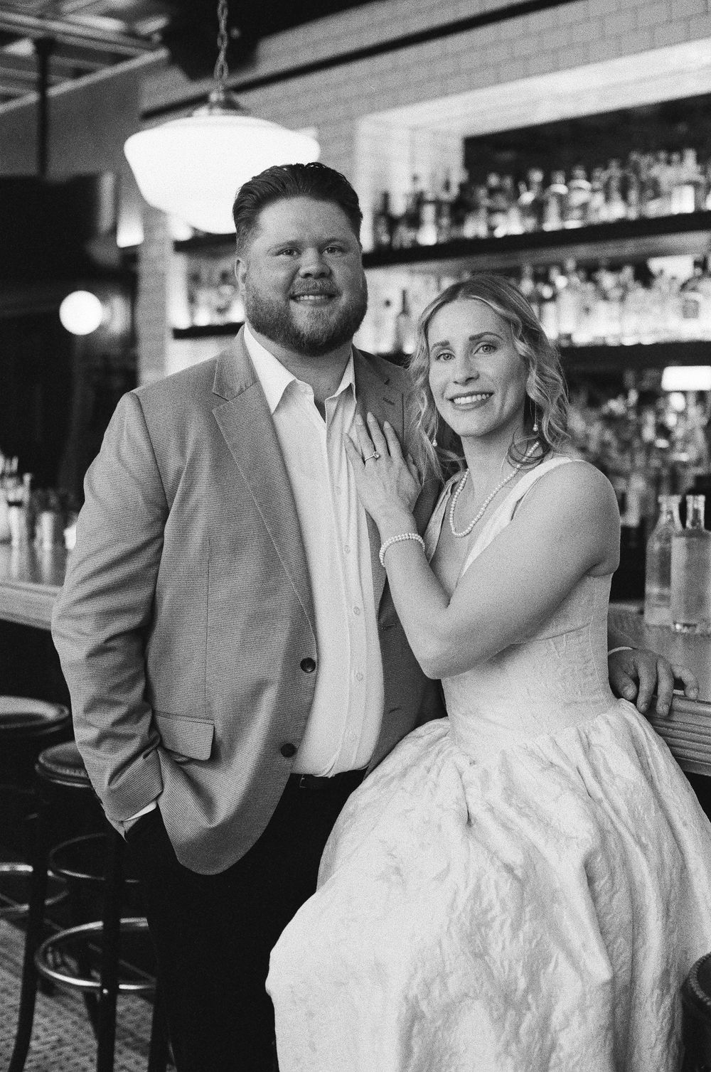 Black and white photo of a smiling couple in formal attire posing together at a bar with shelves of bottles behind them.