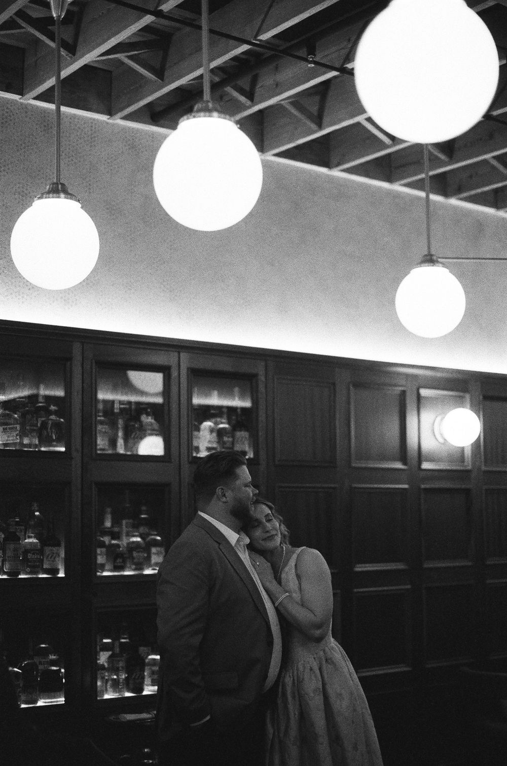 Black and white photo of a couple embracing in a bar with glowing globe pendant lights overhead.