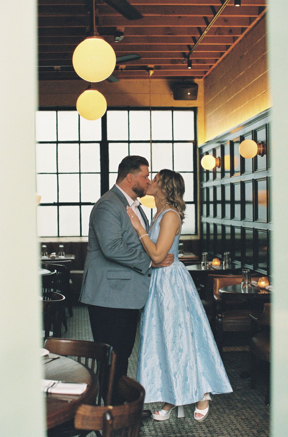 Couple sharing a romantic kiss in an elegant restaurant with large windows and warm pendant lights.