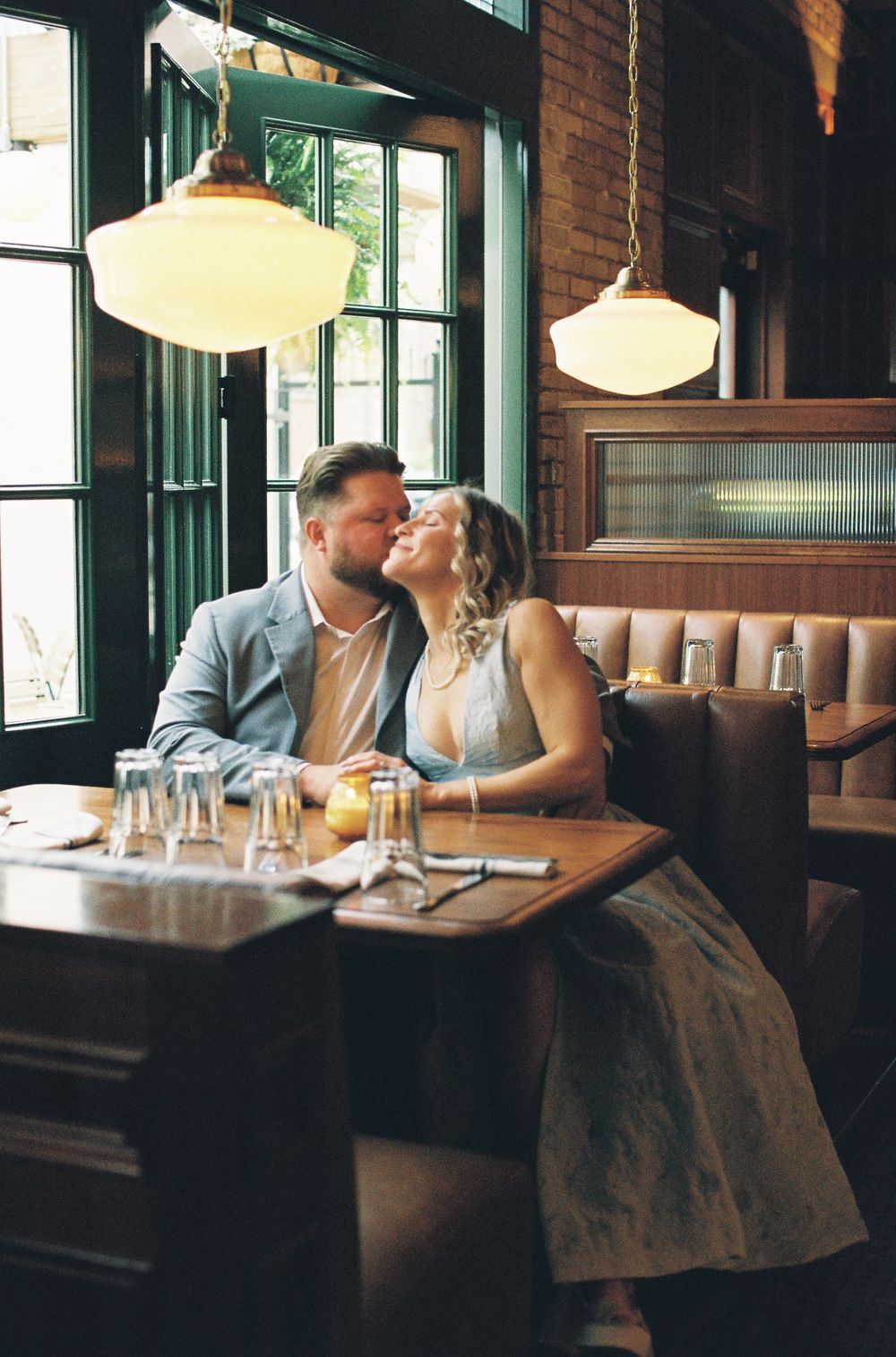 Couple sharing a romantic moment at a restaurant booth, surrounded by warm pendant lights and green windows.