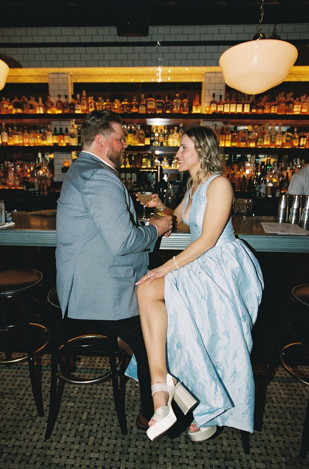Couple in formal attire sharing a romantic moment at an upscale bar with shelves of liquor bottles.