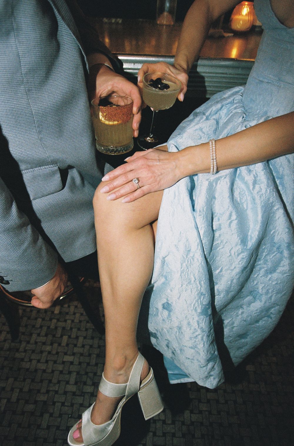 Couple at a formal event holding cocktails, woman in light blue dress with engagement ring and white heeled sandals.