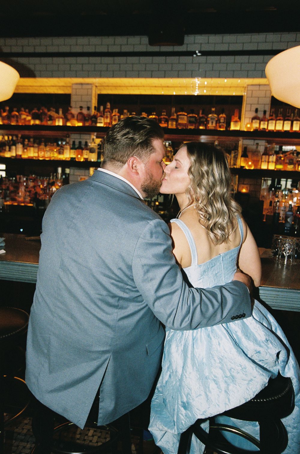 Couple sharing a romantic kiss at a bar with warmly lit liquor shelves in the background.