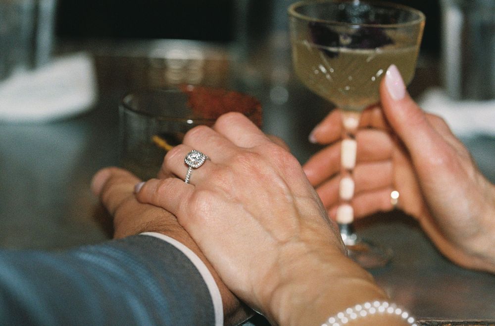 Close-up of hands holding cocktail glasses, woman wearing diamond engagement ring and pearl bracelet at elegant event.