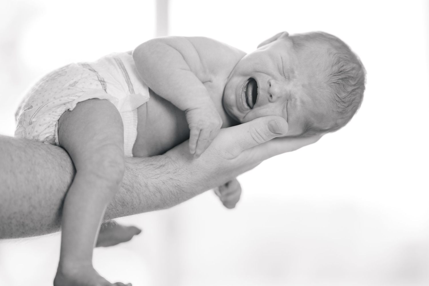 Black and white photo of a newborn baby crying while being cradled in an adult's hands.