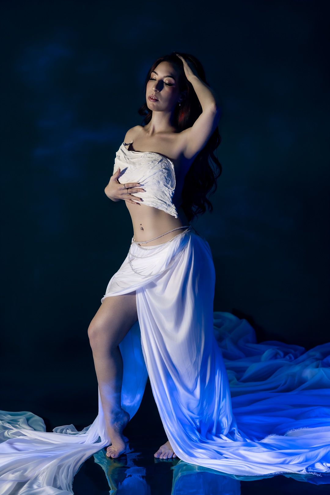 Woman in flowing white dress poses gracefully against dark blue background with dramatic lighting.