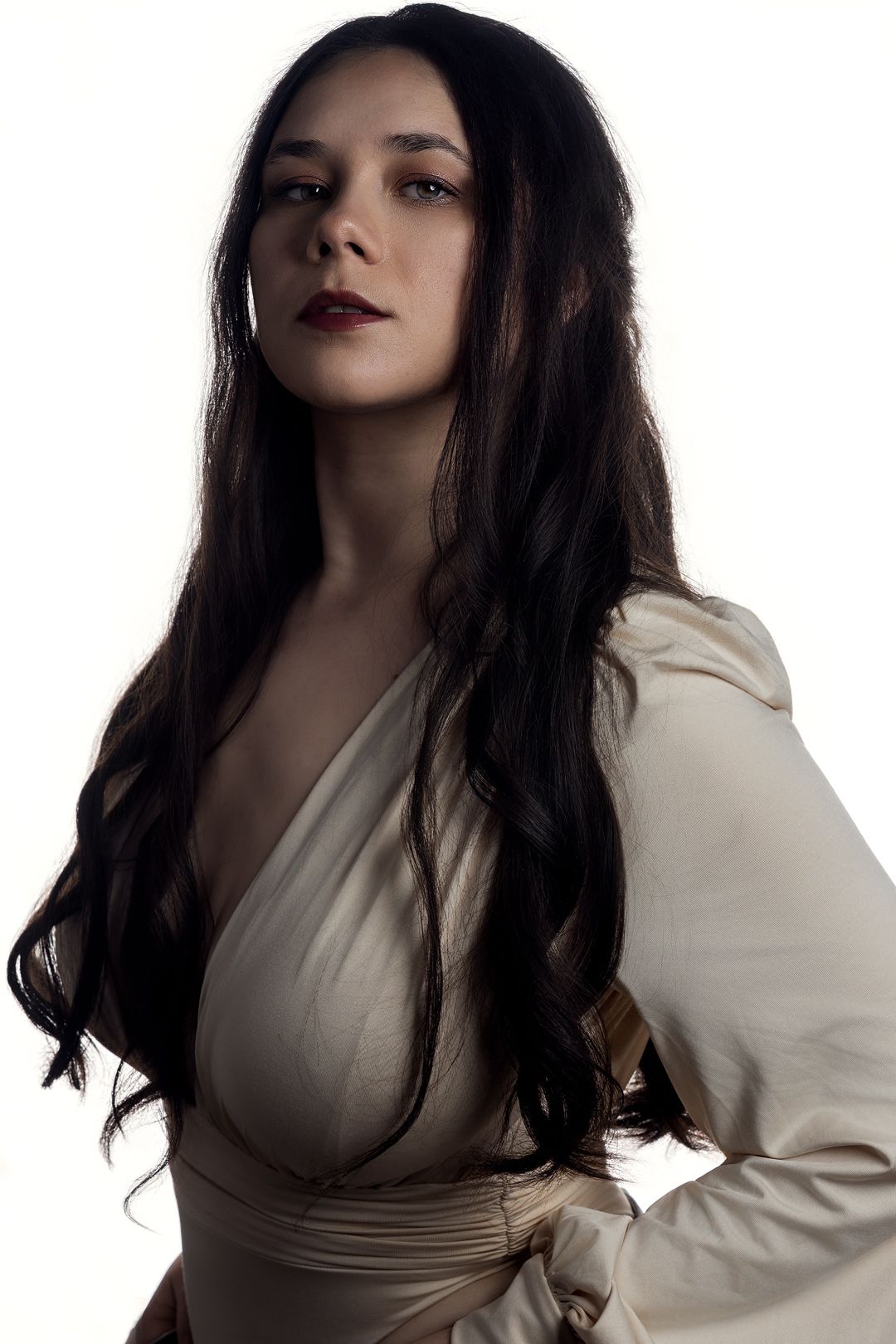 Woman with long dark hair wearing a cream wrap dress poses against a white background.