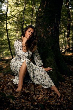 Woman in floral dress sitting against moss-covered tree in forest, surrounded by autumn leaves.