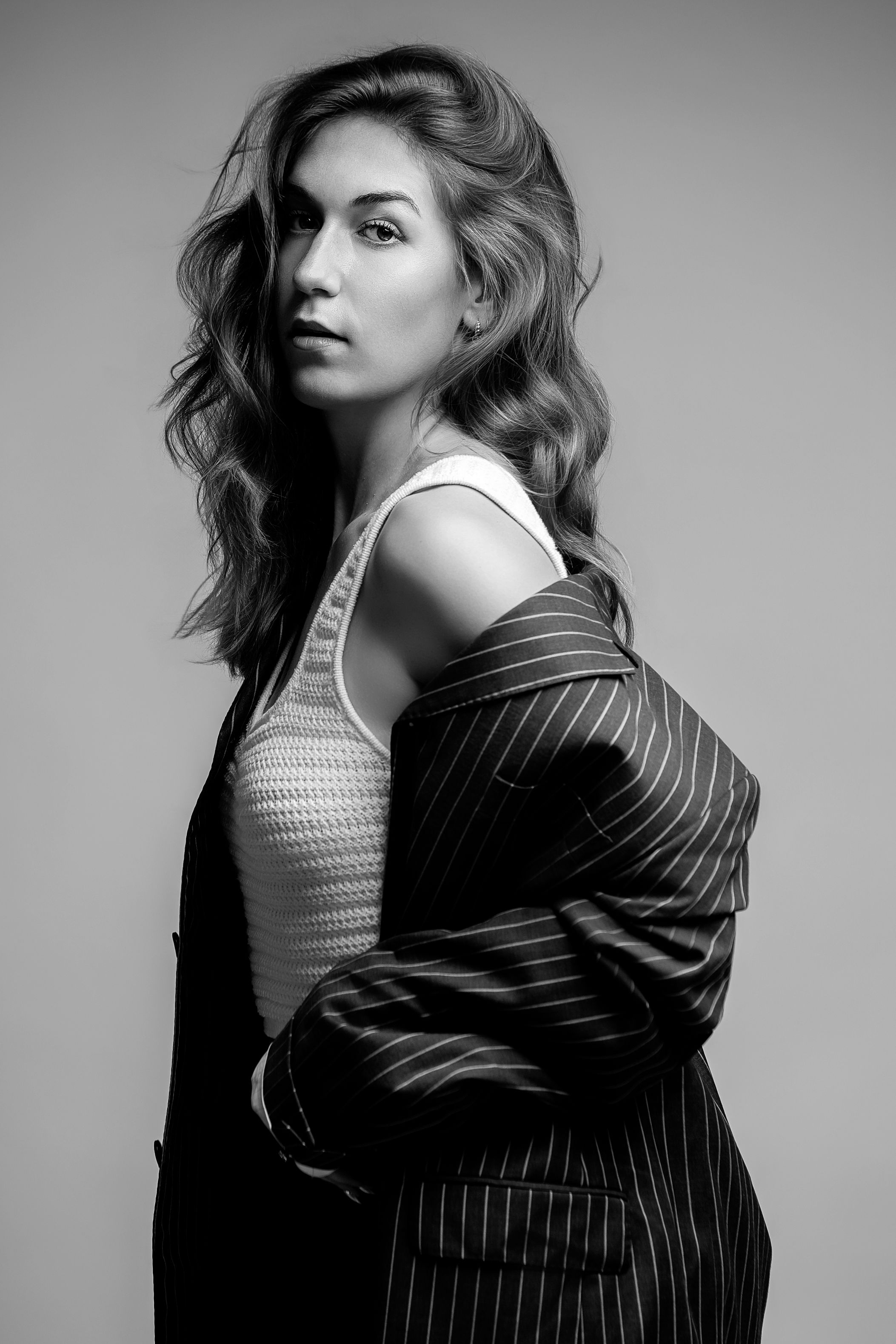 Black and white studio portrait of a woman in a pinstripe blazer and white tank top, posing with wavy hair.