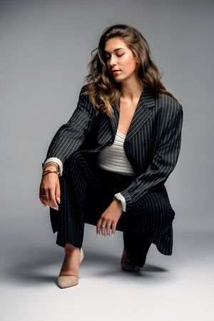 Woman in pinstripe suit crouching gracefully, wearing nude heels and gold bracelets against a gray studio background.