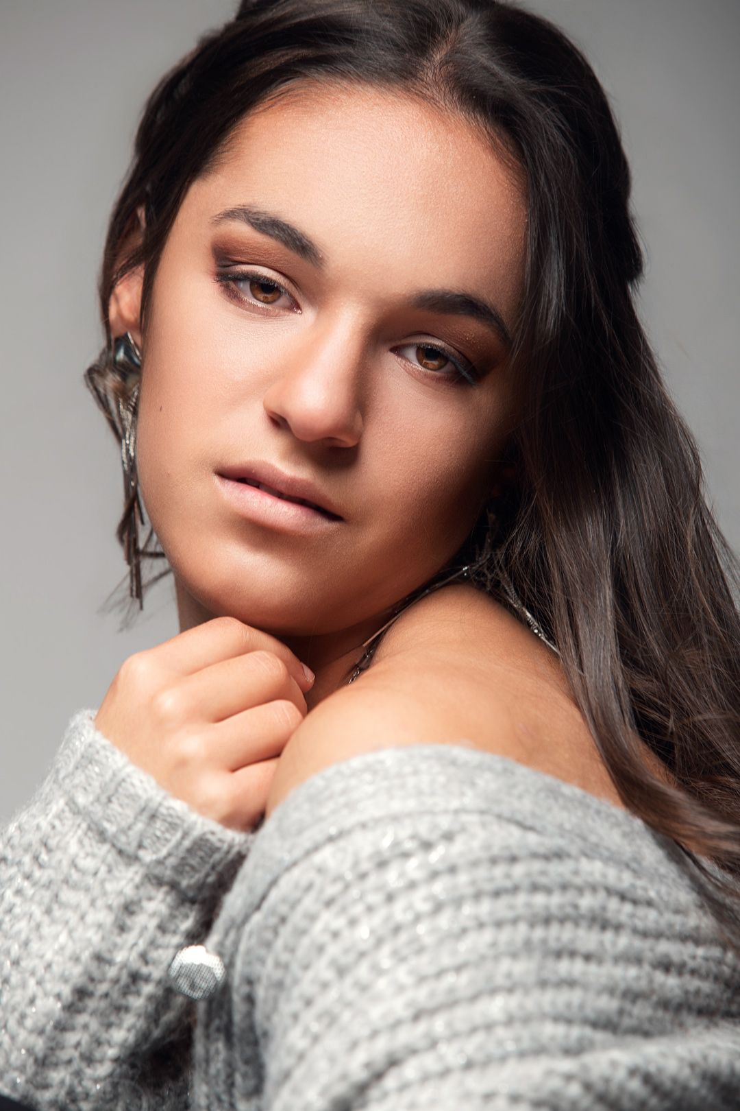 Young woman with dark hair in gray off-shoulder knit sweater, wearing earrings, posing against gray background.
