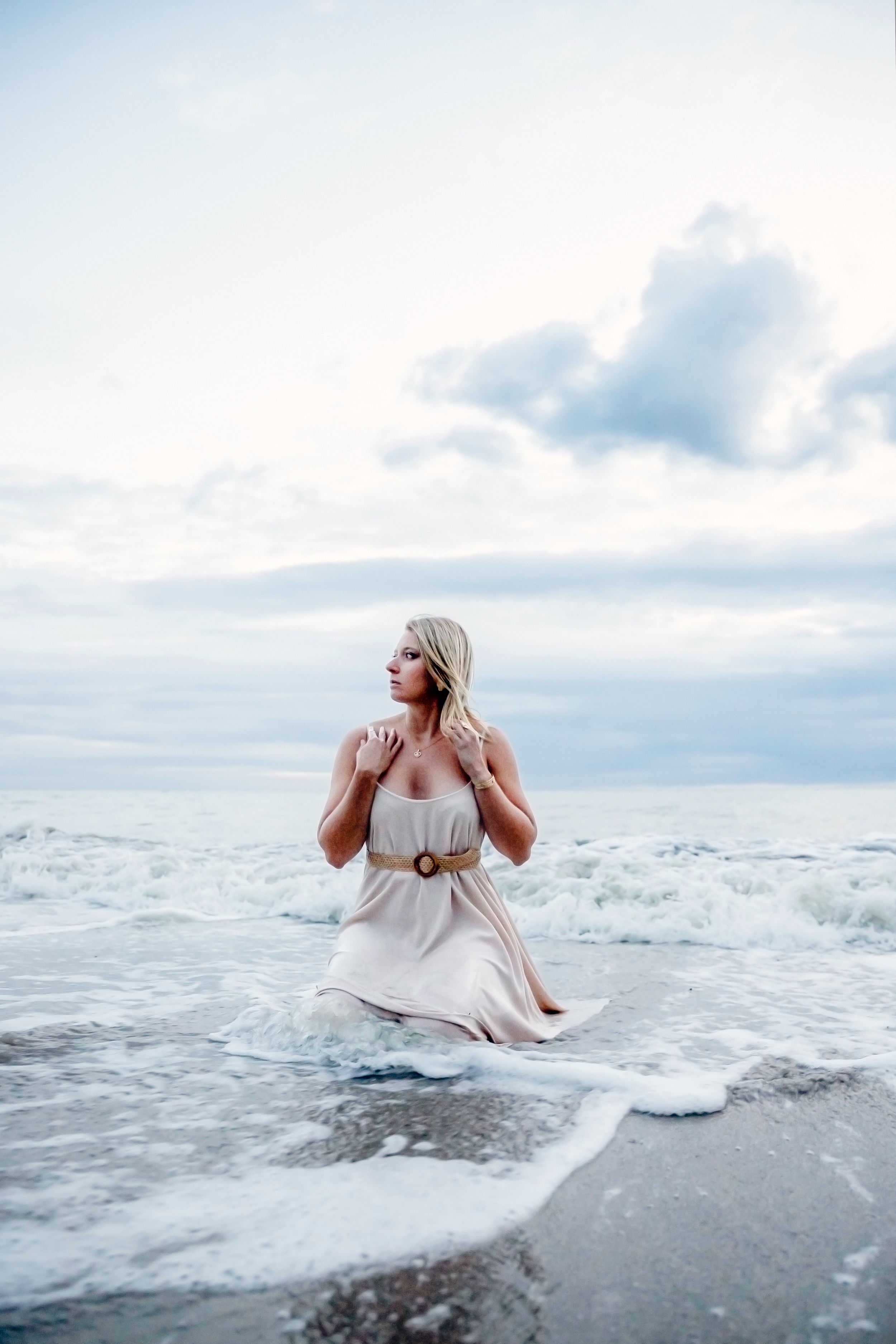 Blonde woman in white belted dress kneeling in ocean waves on cloudy beach.