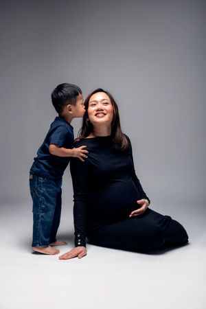 A young boy kisses his pregnant mother on the cheek as she sits on the floor smiling, both dressed in navy blue.