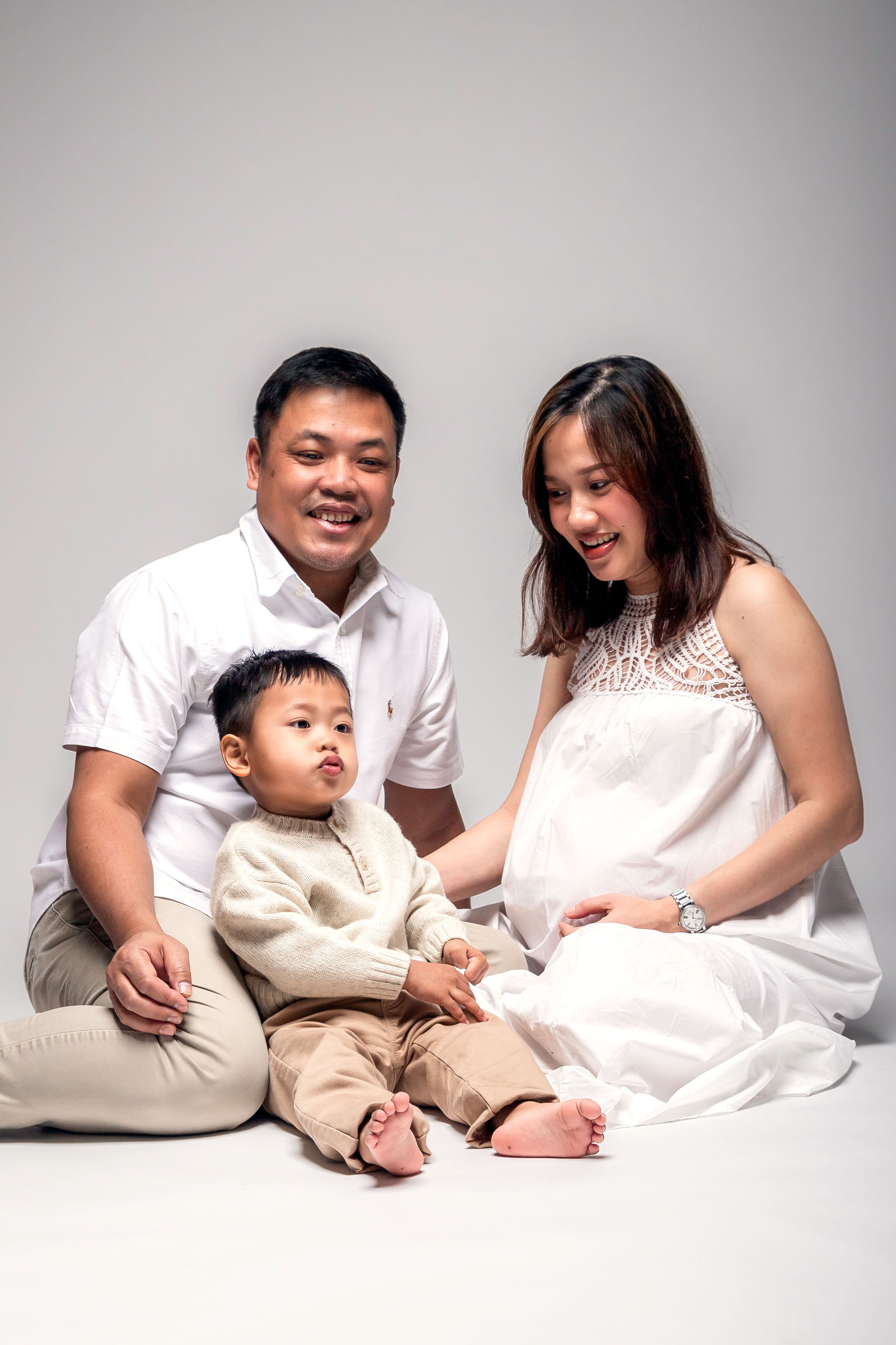 Smiling Asian family with a pregnant mother in white dress, father, and toddler son posing together on a white background.