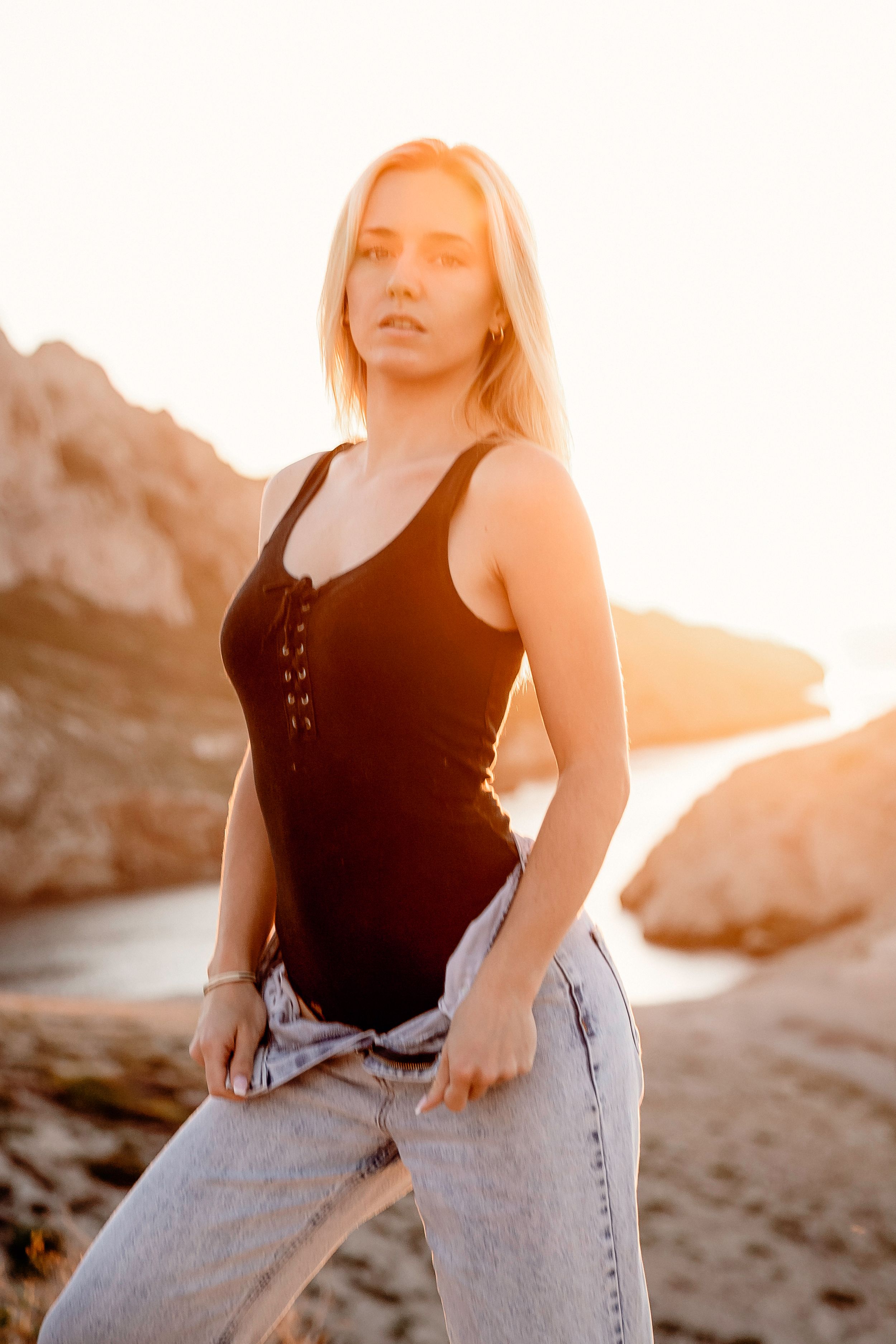 Blonde woman in black tank top and light wash jeans poses on rocky beach at golden sunset.