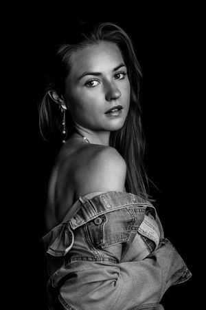 Black and white portrait of a young woman in an off-shoulder denim jacket, looking over her shoulder against dark background.