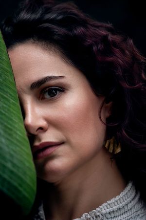 Close-up portrait of a woman with curly dark hair and gold earrings, partially hidden behind a green tropical leaf.