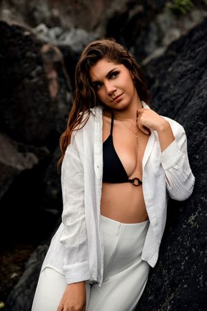 Young woman in white open shirt and black bikini top posing against dark rocks outdoors.