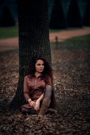 A woman in a brown dress and boots sits against a large tree trunk on a bed of fallen autumn leaves in a park.