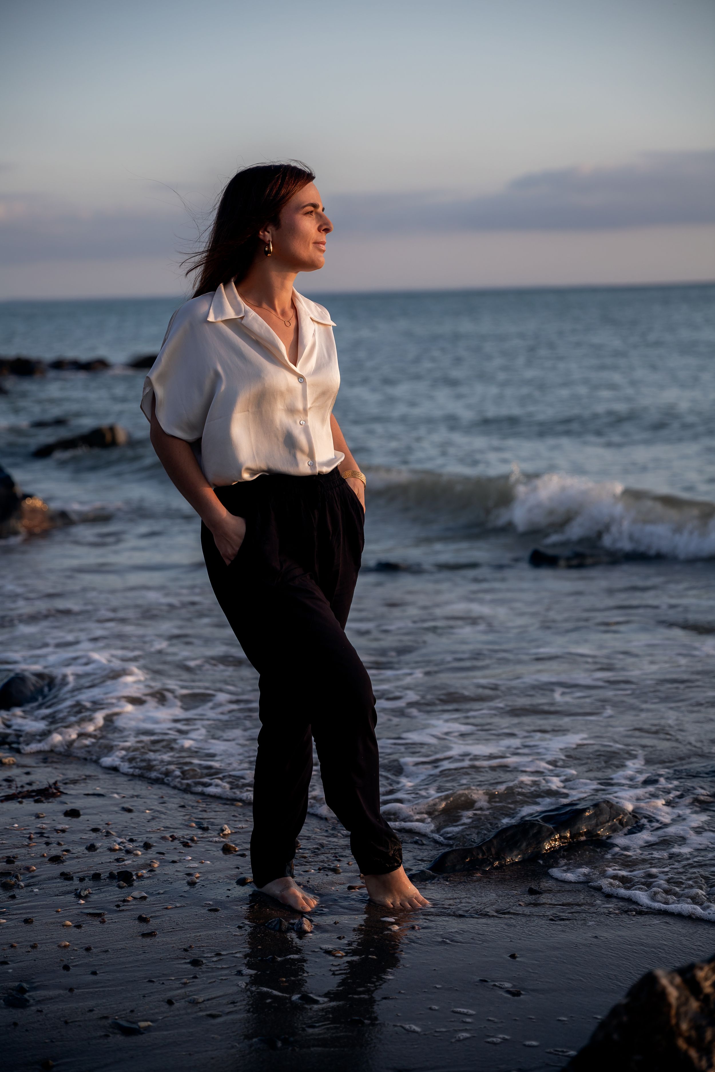 Woman in white shirt and black pants standing barefoot on a rocky beach at sunset with ocean waves behind her.