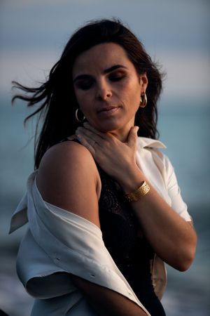 A woman with dark hair poses by the sea, wearing a white shirt, black dress, gold hoop earrings, and a gold cuff bracelet.