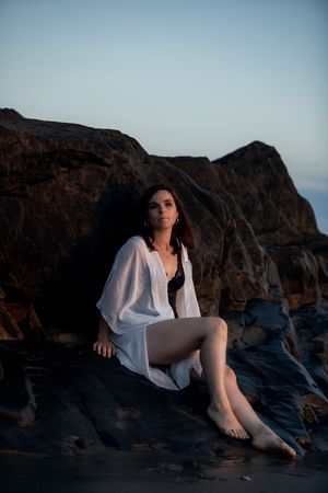 Woman in white shirt sitting on coastal rocks at dusk, posing barefoot against dramatic cliffs.