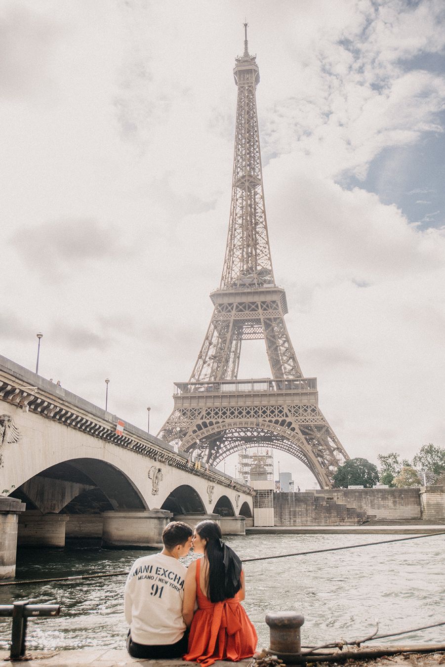 Couple sitting by the Seine River sharing a romantic moment with the iconic Eiffel Tower in Paris, France behind them.