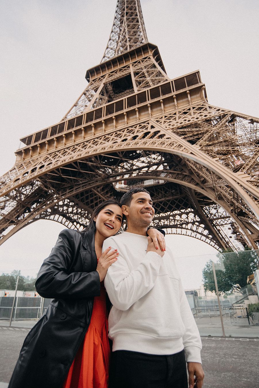 Happy couple embracing in front of the Eiffel Tower in Paris, woman in red dress and black jacket.