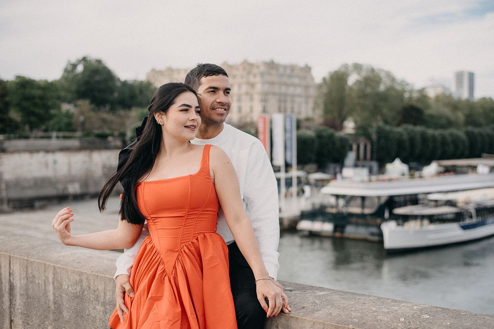 A couple poses romantically by the Seine River in Paris, woman in orange dress, man in white shirt.