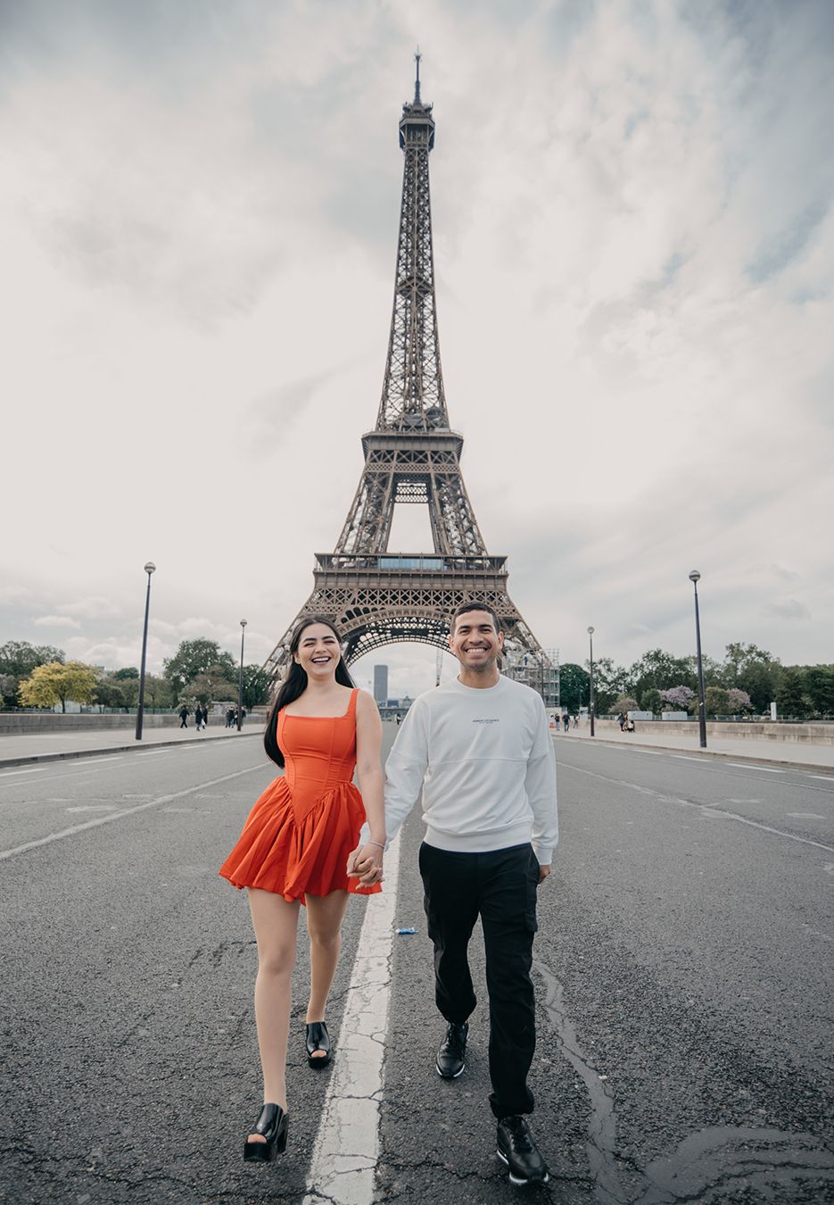Couple holding hands walking in front of the Eiffel Tower in Paris, woman in red dress, man in white sweater.