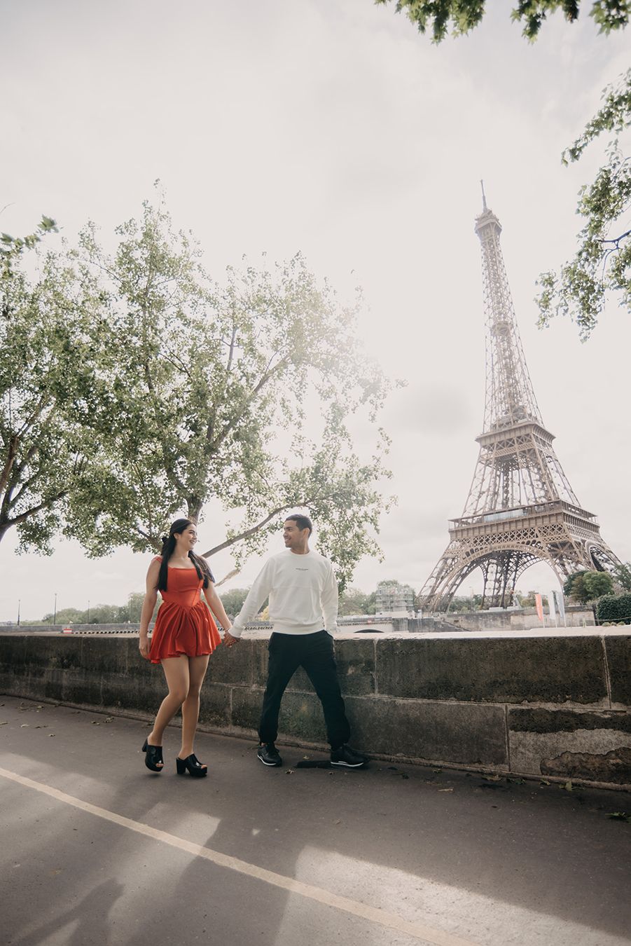 Couple holding hands walking near the Eiffel Tower in Paris, woman in red dress, man in white sweater.