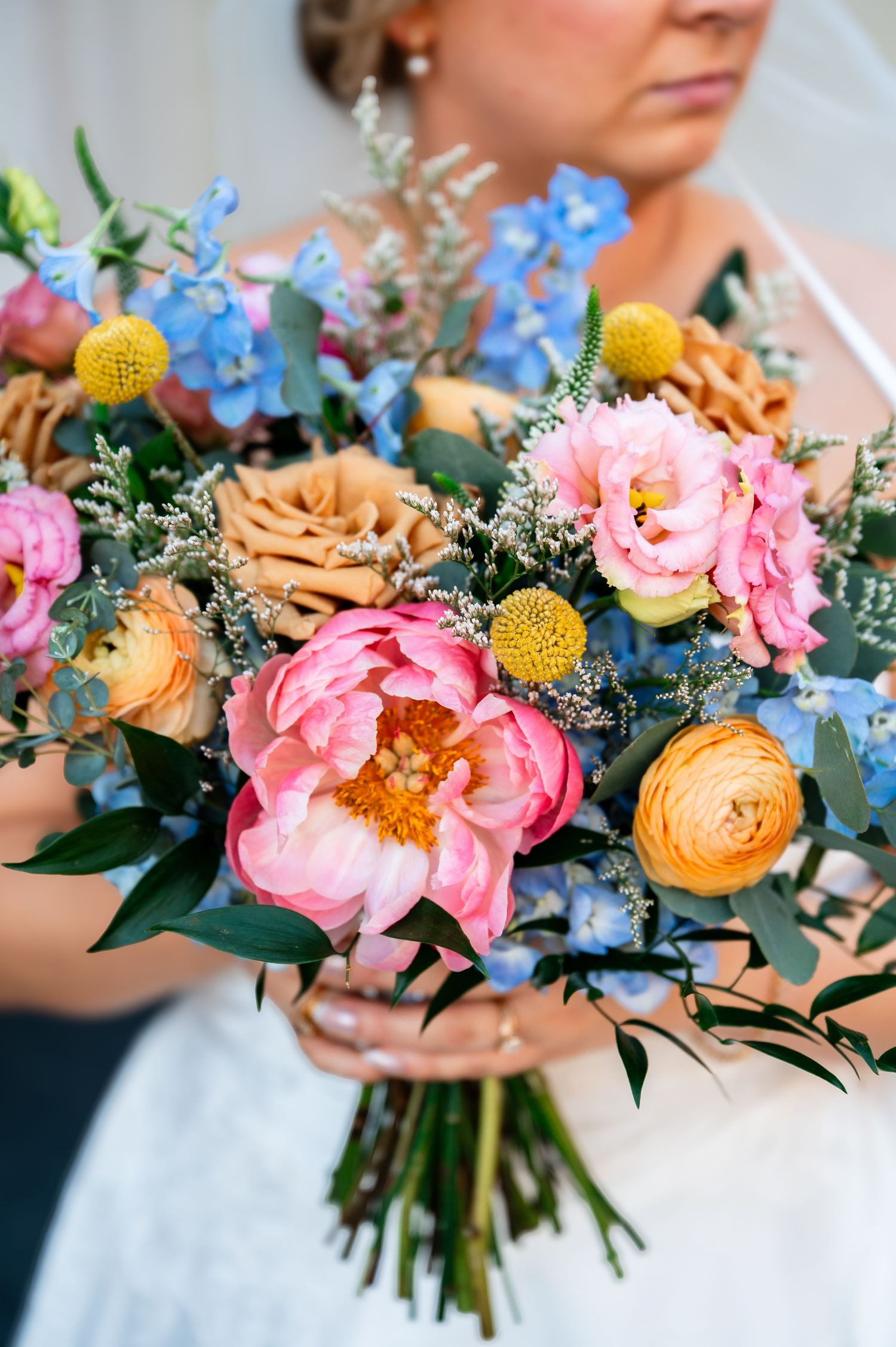 Bride holding a vibrant wedding bouquet with pink peonies, blue delphinium, orange ranunculus, and yellow craspedia flowers.