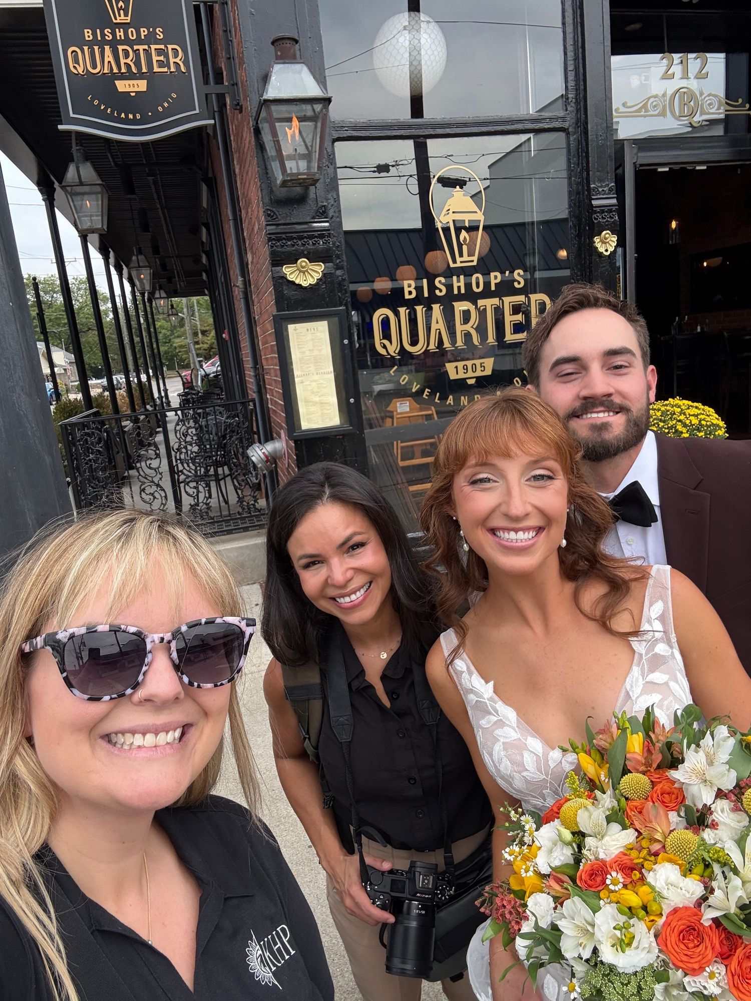 Wedding party selfie outside Bishop's Quarter restaurant, bride holding colorful bouquet with groom in tuxedo.