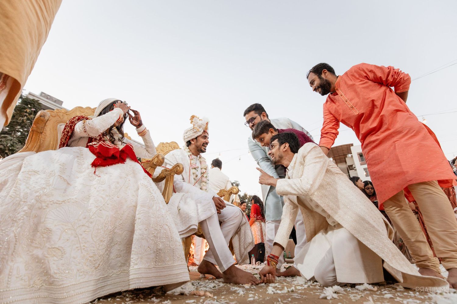 Indian wedding ceremony with groom in white sherwani surrounded by groomsmen in coral kurtas outdoors.