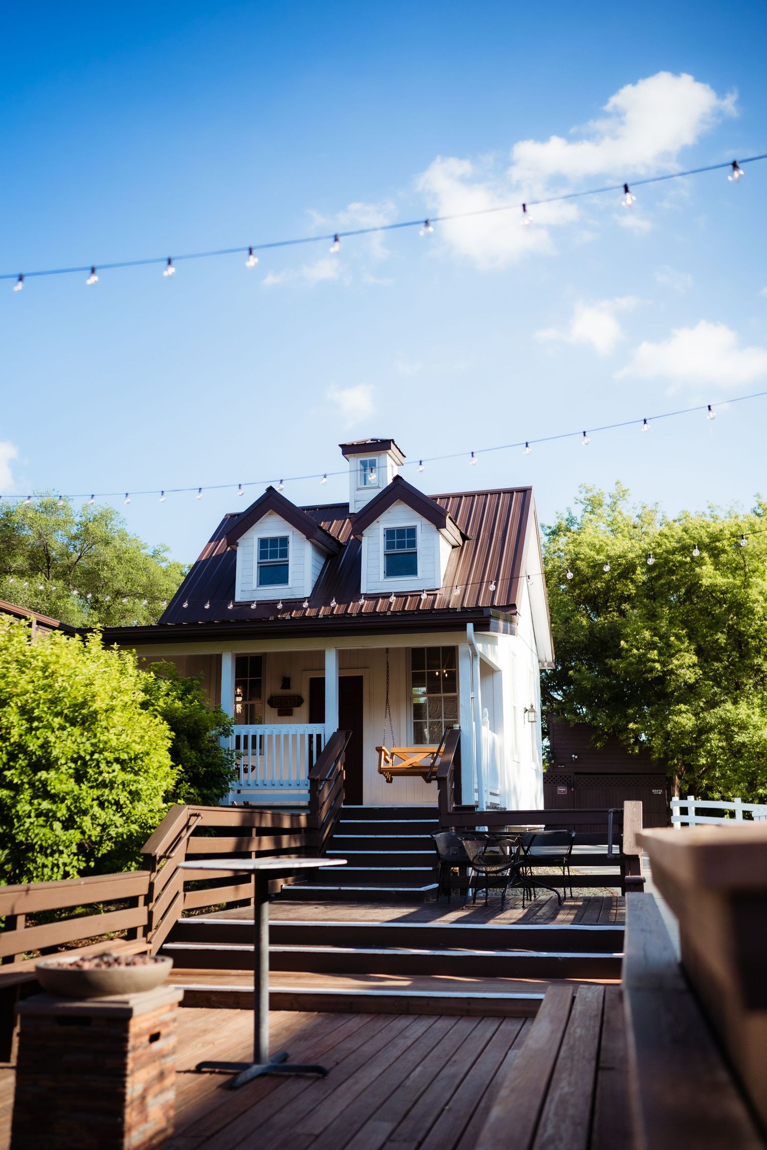 Charming white cottage with brown metal roof, string lights, and wooden deck patio surrounded by lush green trees under blue sky.