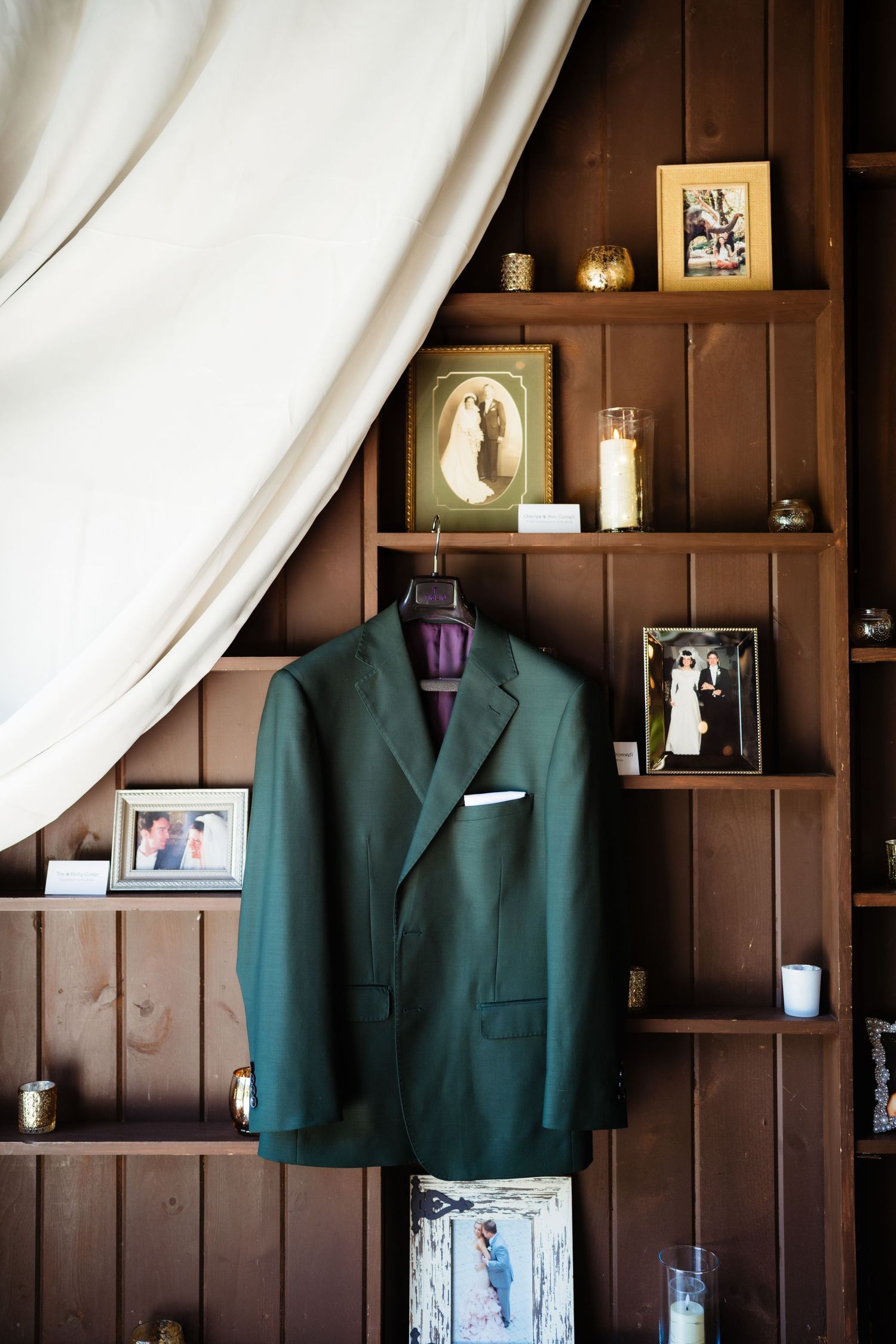 Green suit jacket hanging on wooden shelving unit adorned with framed wedding photos, candles, and white curtain drape.