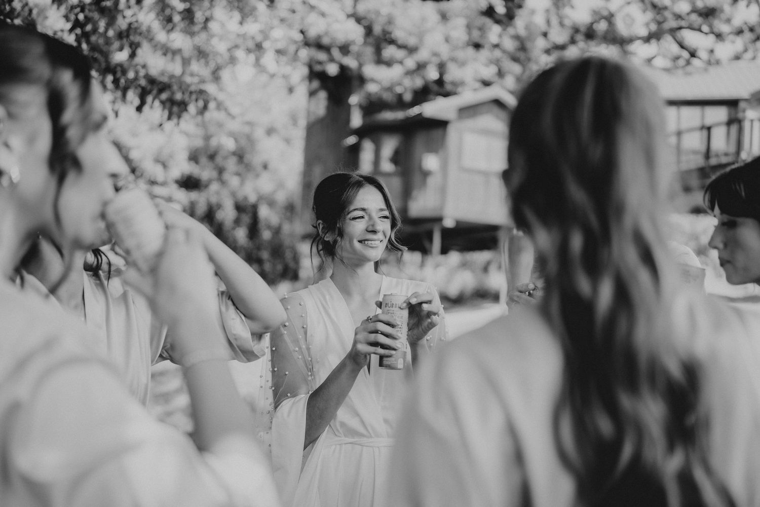 Black and white photo of a smiling bride toasting with bridesmaids outdoors at a wedding celebration.